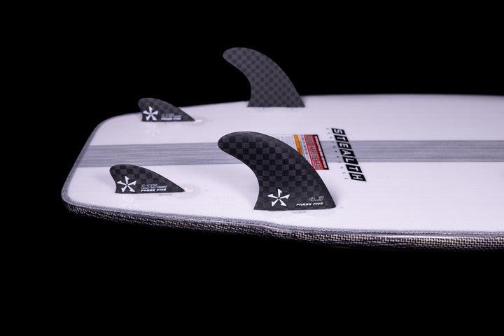 Close-up of a 2025 Phase 5 Swell Stealth surfboard with black carbon fiber fins on a white board against a black background.