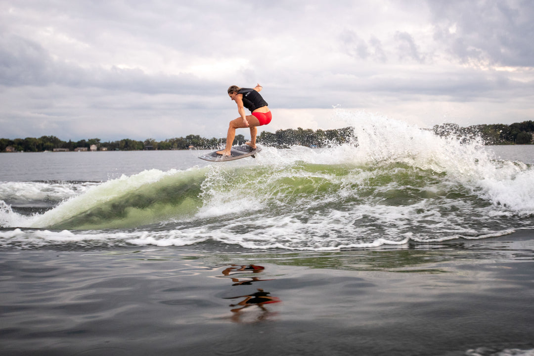 A person surfing on the 2025 Phase 5 Swell Stealth Surf Board on a wave, with a cloudy sky and shoreline in the background.