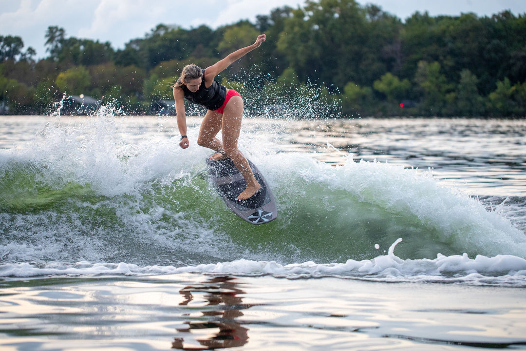 A woman surfing on a 2025 Phase 5 Swell Stealth Surf Board, riding a wave with a lush green shoreline in the background.