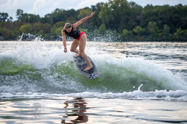 A woman surfing on a 2025 Phase 5 Swell Stealth Surf Board, riding a wave with a lush green shoreline in the background.