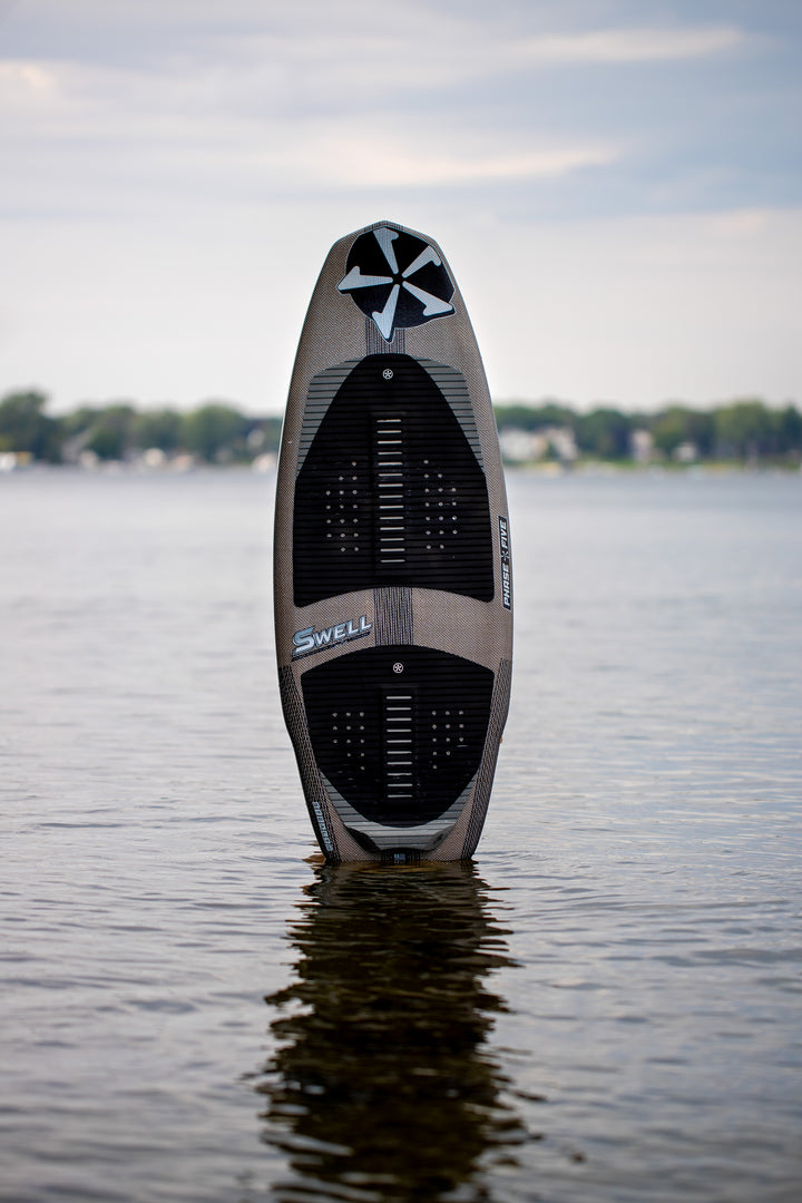 2025 Phase 5 Swell Stealth surfboard standing upright in calm water with a blurred shoreline in the background.