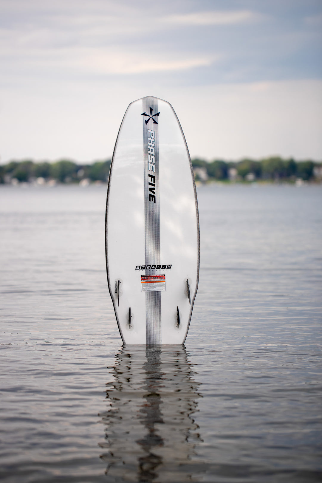 A white Phase 5 Swell Stealth surfboard standing upright in calm water, with a blurred shoreline and cloudy sky in the background.