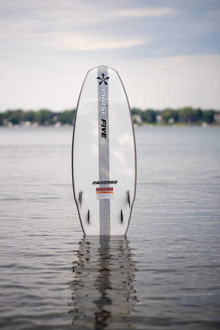 A white Phase 5 Swell Stealth surfboard standing upright in calm water, with a blurred shoreline and cloudy sky in the background.