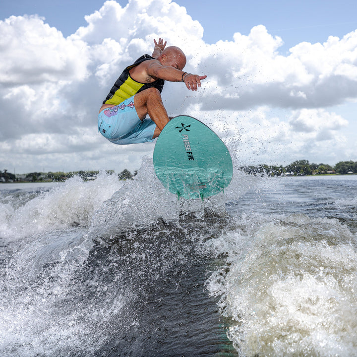 A surfer performs a jump on the 2026 Phase Five Trident V3 board, showcasing agility on water with a vibrant blue sky backdrop.