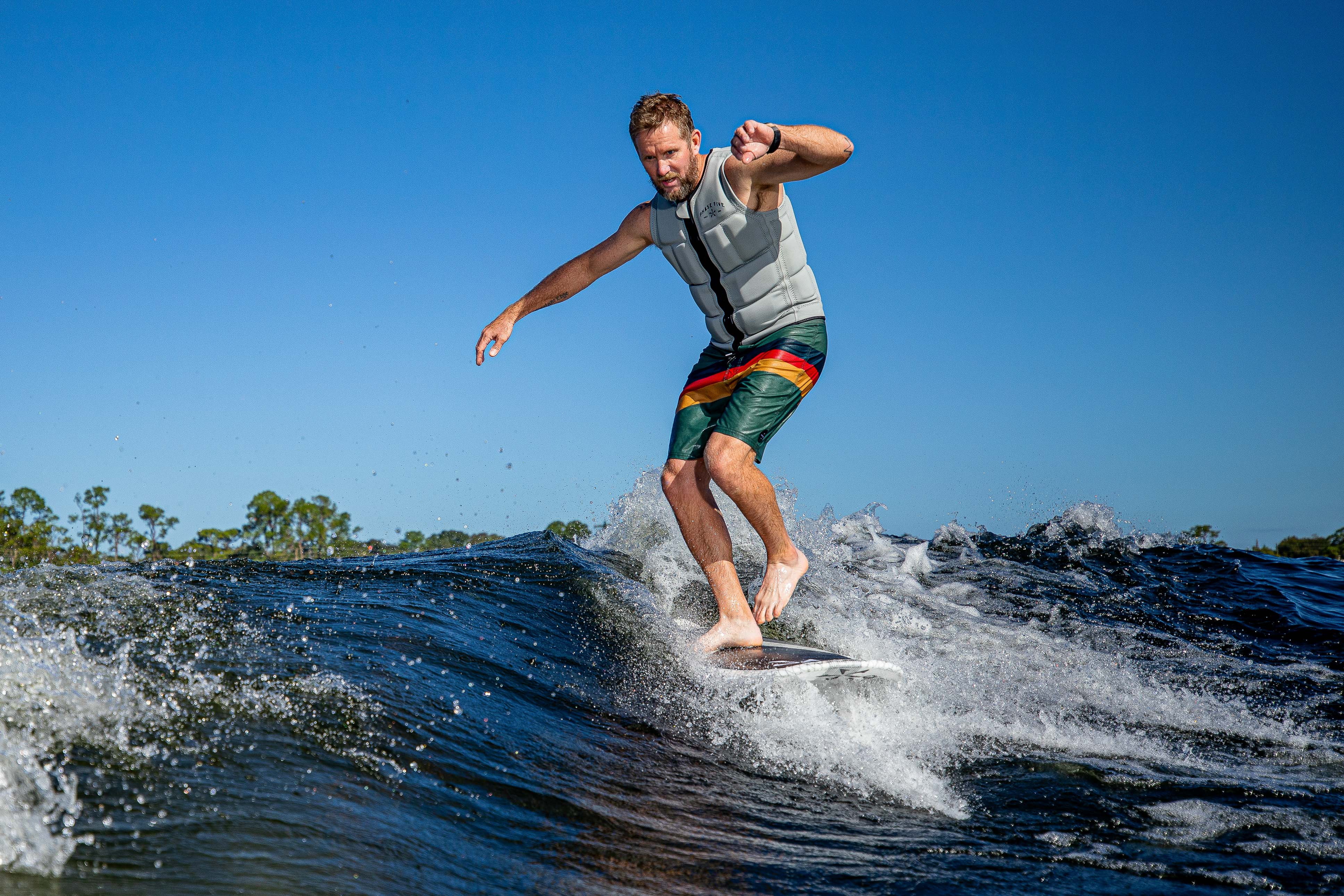 Man surfing on the 2025 Phase 5 Zilla Surf Board, balancing on a wave with a clear blue sky background.