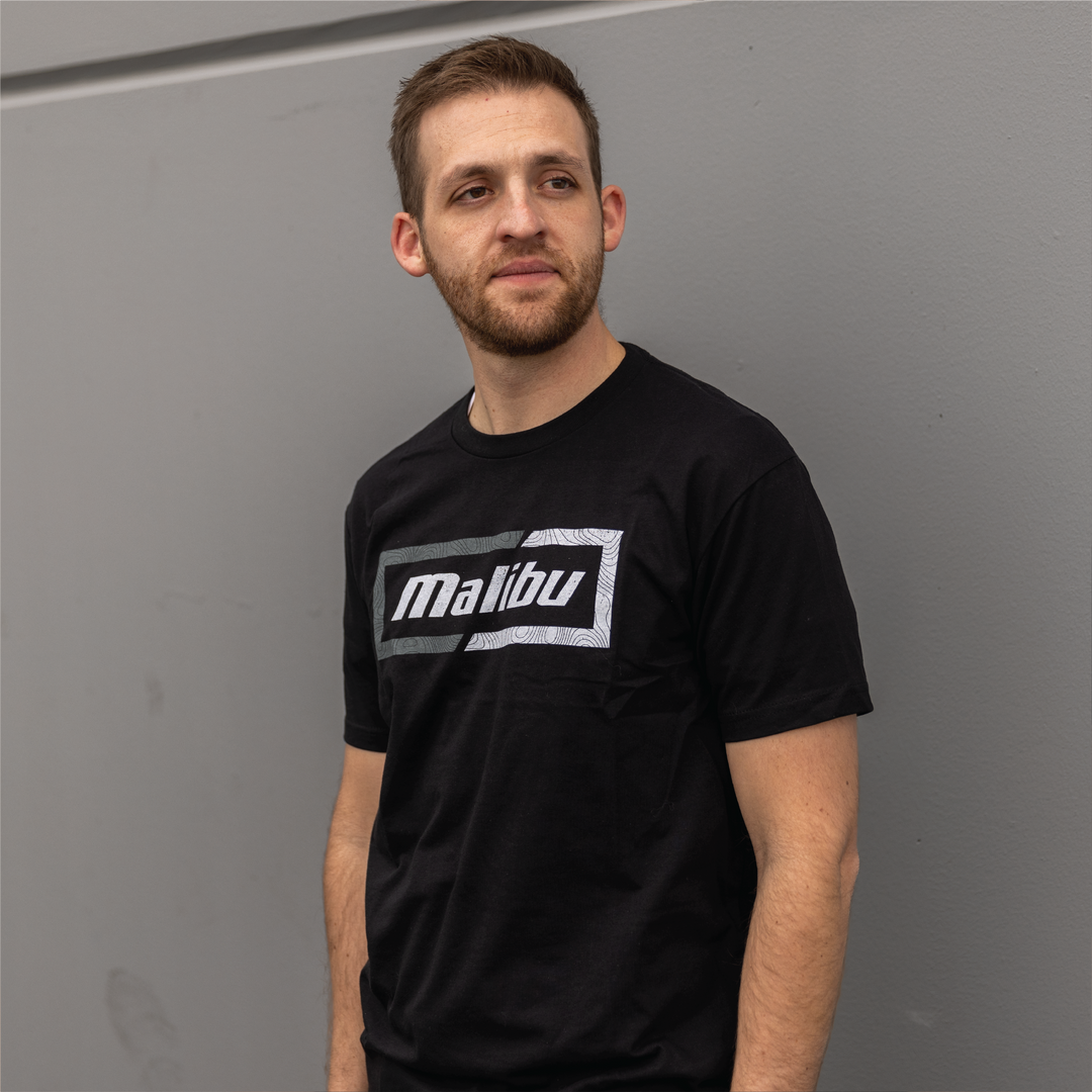 Man wearing a black Malibu Topo Tee with a graphic logo on the front, standing against a gray wall.