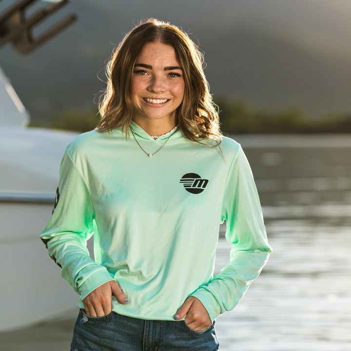 Young woman smiling outdoors in a green shaded shredder sun hoodie by the water, with a boat and mountains in the background.
