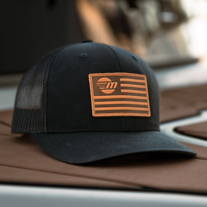 Black trucker hat with a tan leather patch featuring a stylized American flag and "FM" logo.