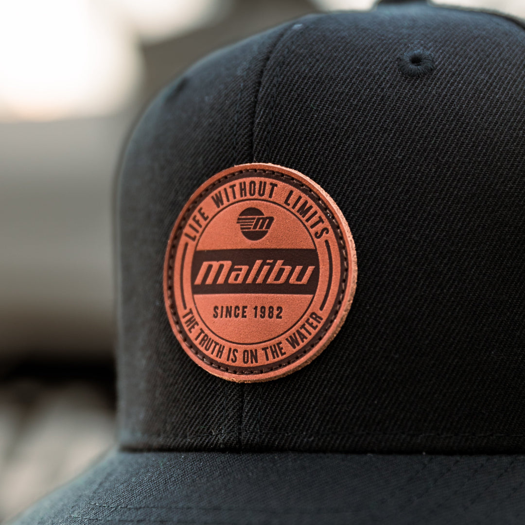 Close-up of a black Malibu hat with a round, pink patch that reads "Life Without Limits" and "Since 1982," on a textured fabric.