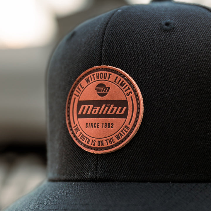 Close-up of a black Malibu hat with a round, pink patch that reads "Life Without Limits" and "Since 1982," on a textured fabric.