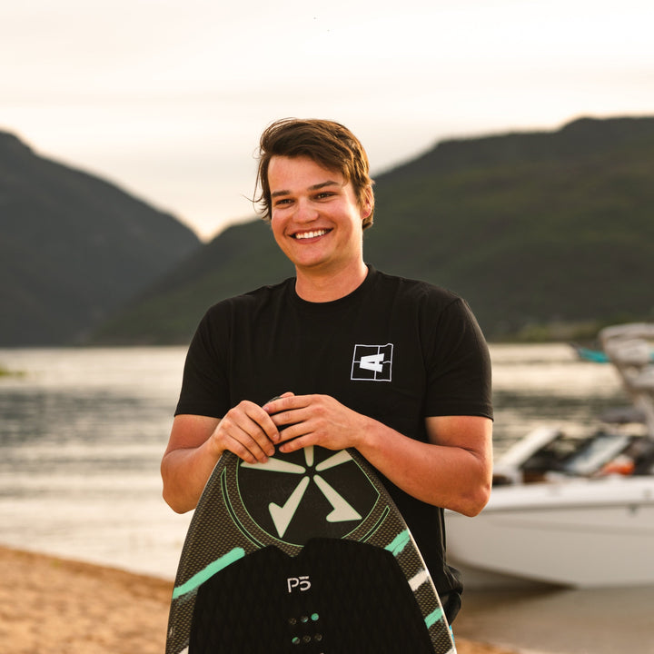 Young man smiling outdoors wearing a black Axis Logo Tee, holding a wakeboard near a lake with mountains in the background.