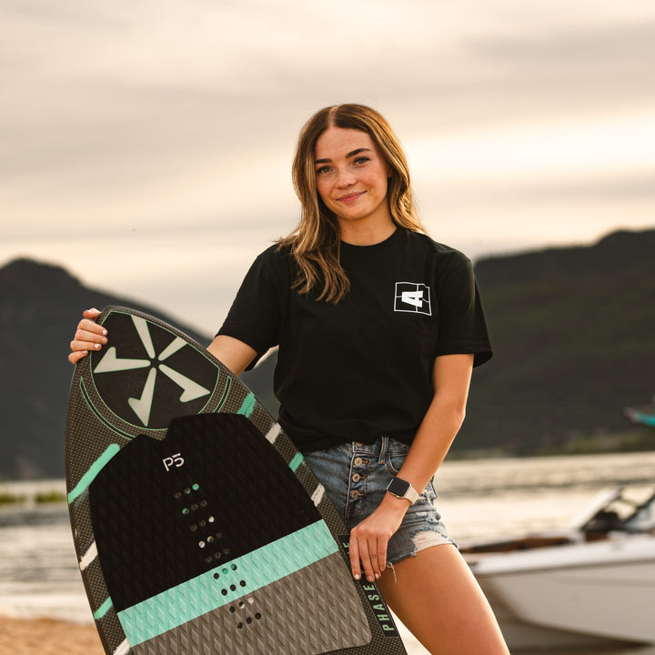 Young woman holding a wakeboard outdoors, wearing a black Axis Logo Tee, denim shorts, and a smartwatch at sunset.