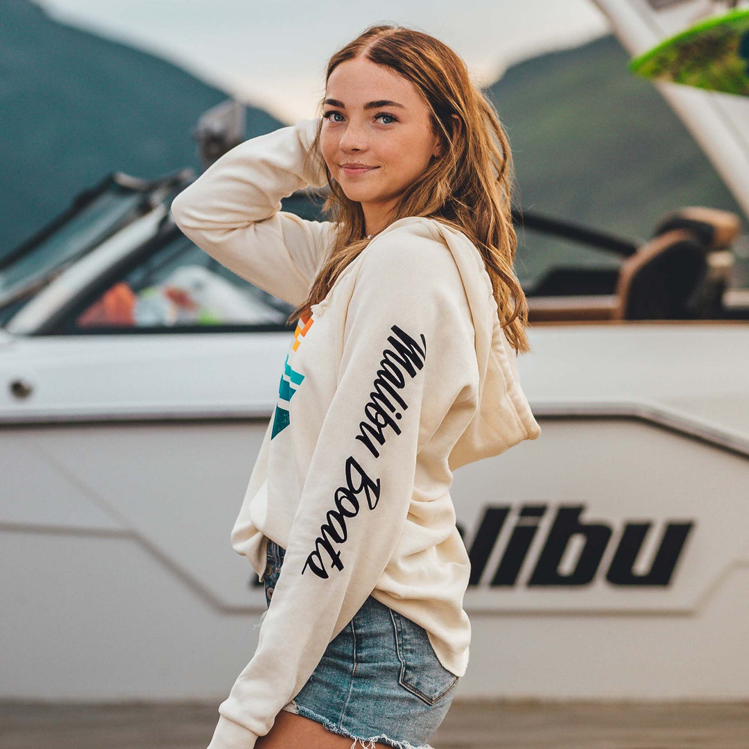 Woman wearing a Sunsetter Pullover hoodie with colorful logo, standing outdoors near a boat, smiling with her hand behind her head.