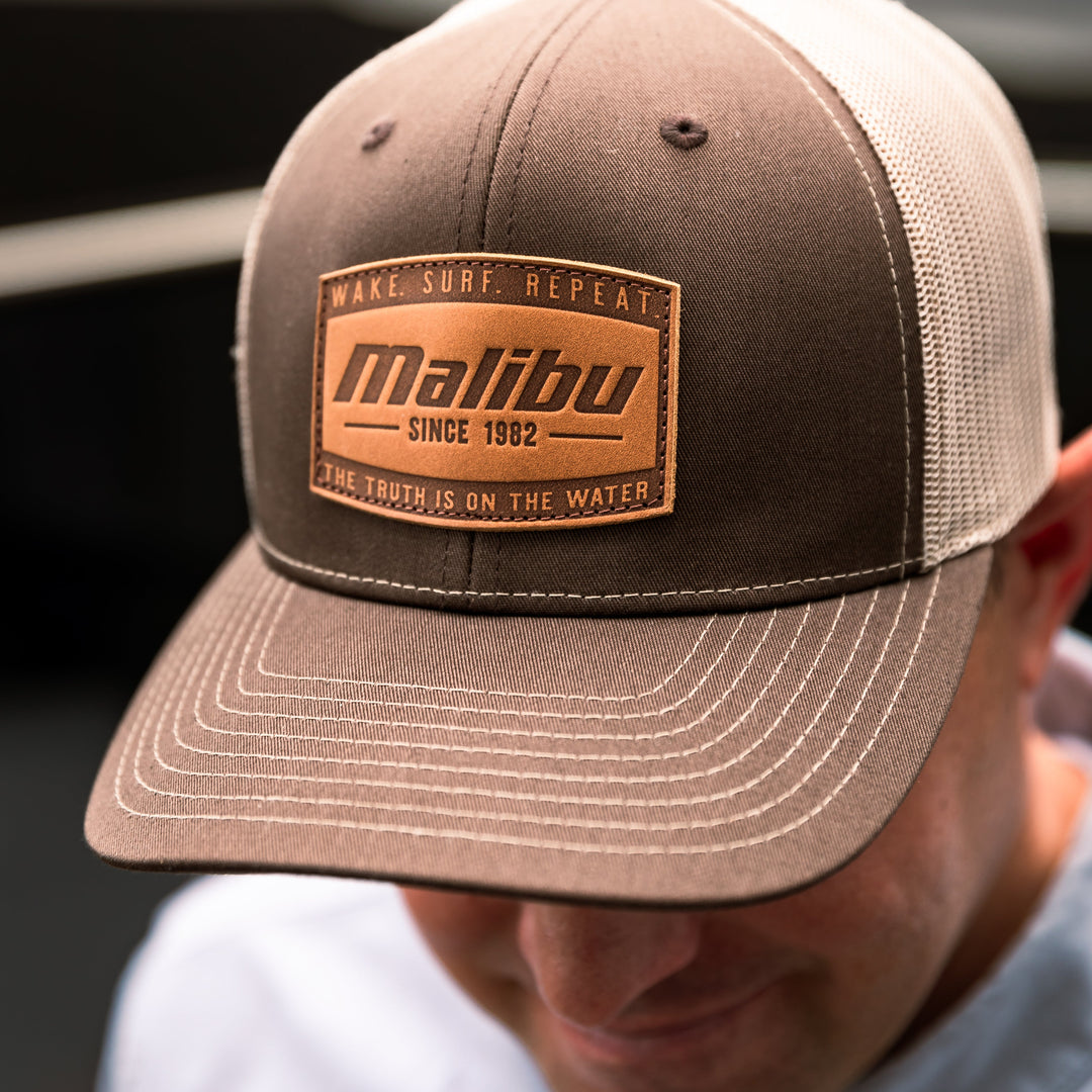 Close-up of a brown and beige trucker-style ranger hat with a patch reading "Malibu" and "Since 1982," worn by a person with a white shirt.