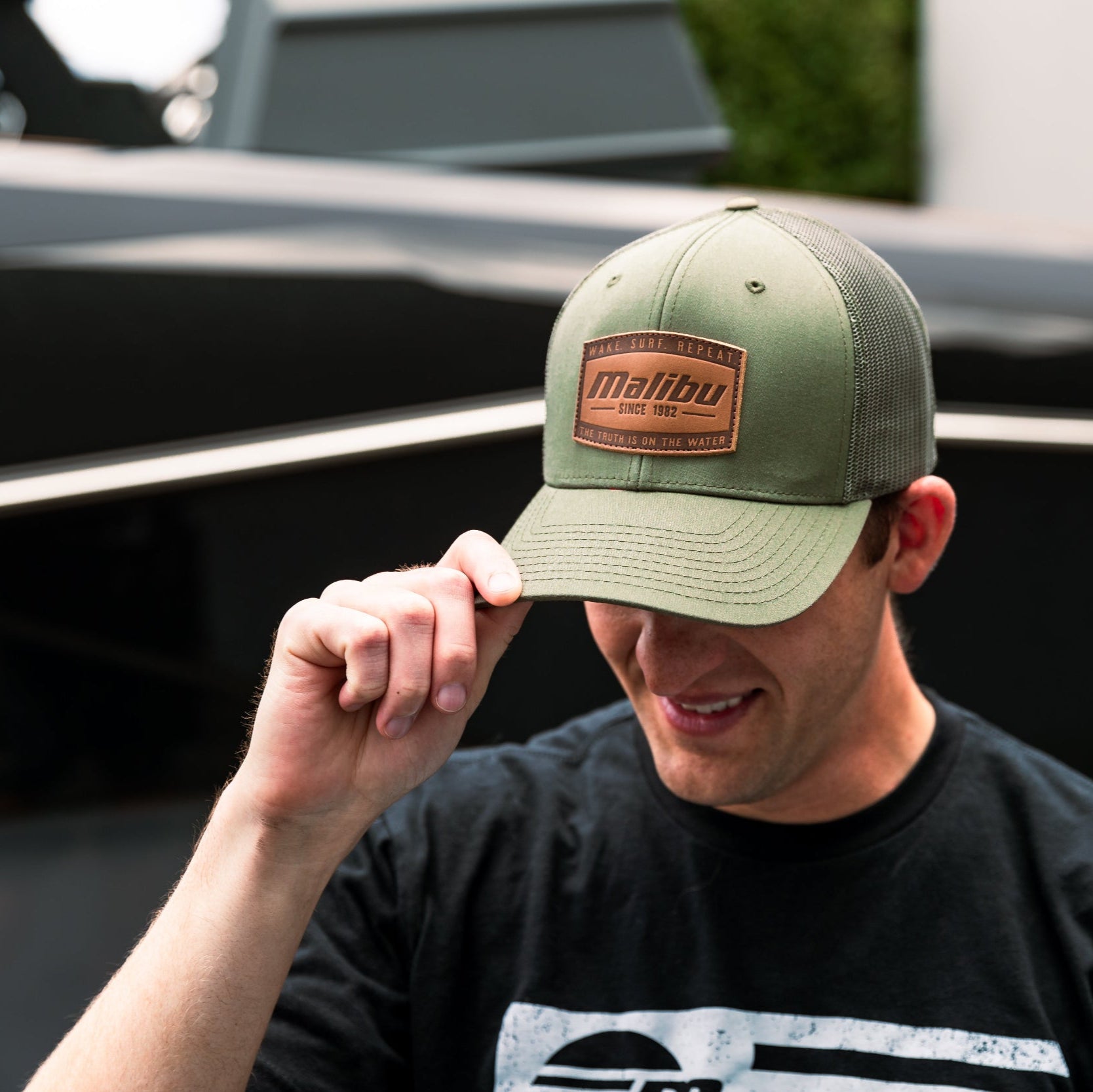 Man wearing a green ranger hat with a brown patch, adjusting the brim outdoors near a boat.