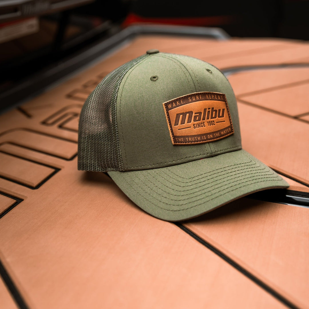 Olive green trucker-style ranger hat with a brown patch reading "Malibu" and "Since 1982" placed on a wooden surface.