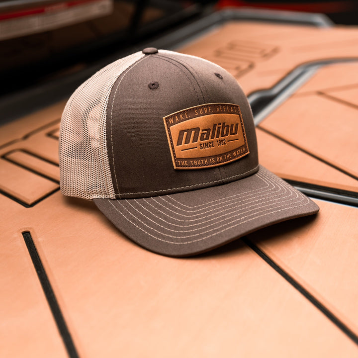 Gray and beige trucker-style ranger hat with a leather patch reading "Malibu" and surf-inspired slogans, placed on a wooden surface.