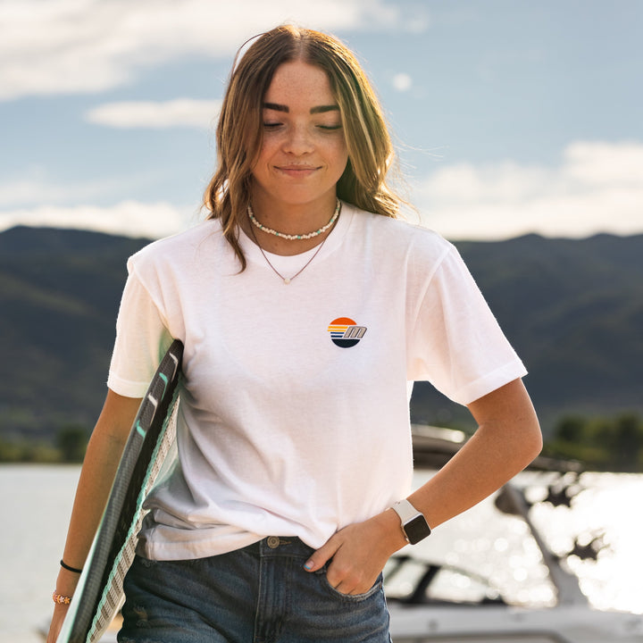 Young woman wearing a white Malibu Monochrome Tee with a small sunset logo, outdoors near water and mountains.