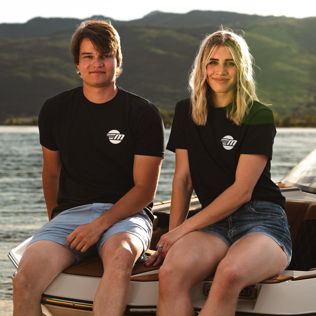 Two young adults sit on a boat wearing black Malibu Monochrome Tees with white logo, enjoying a lakeside view at sunset.
