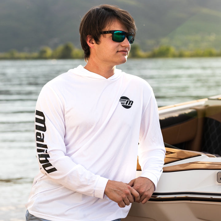 Man wearing a white Shaded Shredder Sun Hoodie with black logo and text, standing near a boat on a lake with mountains in the background.