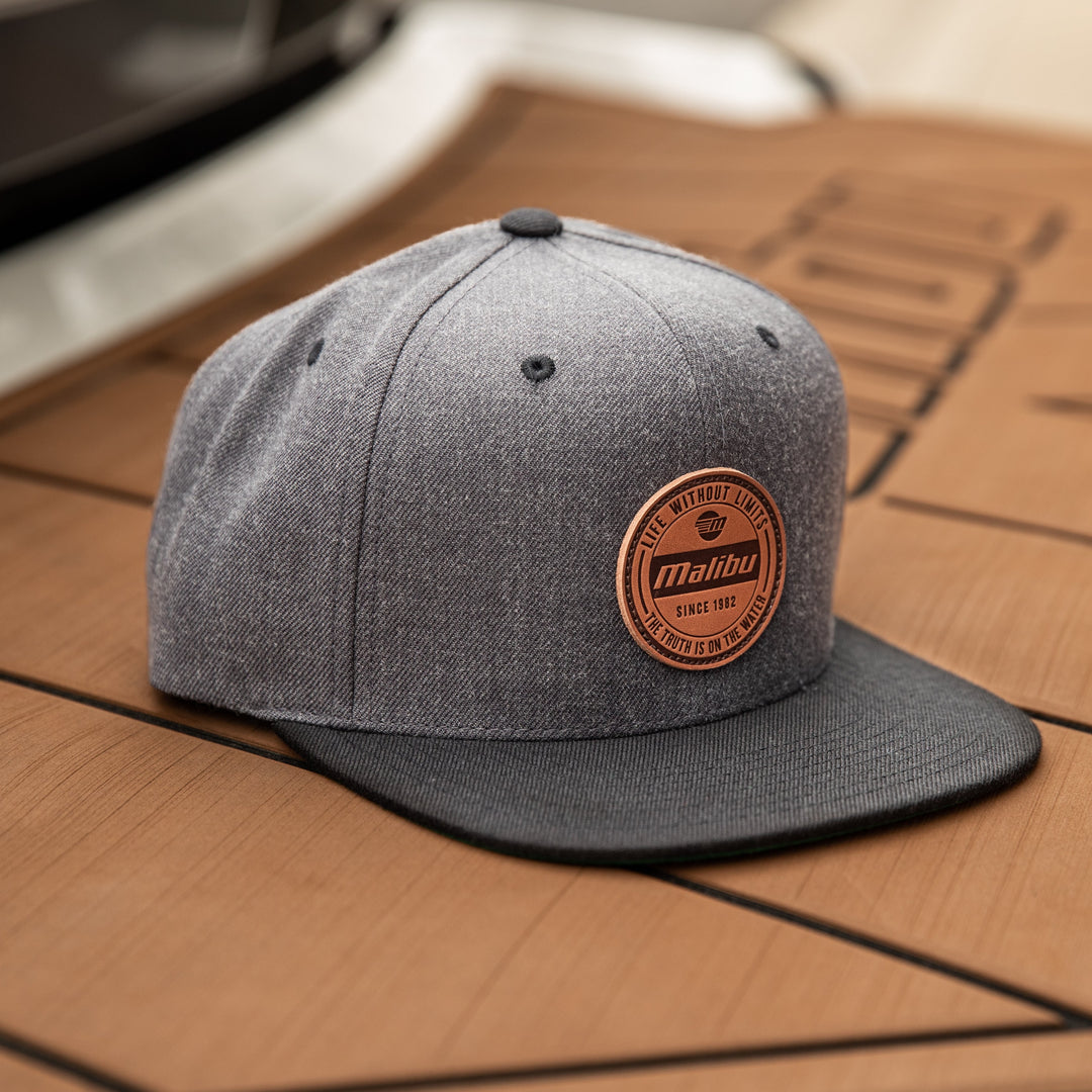 Gray snapback hat with a circular leather patch reading "Malibu, Since 1982, Life Without Limits, The Truth Is On The Water" on a wooden surface.