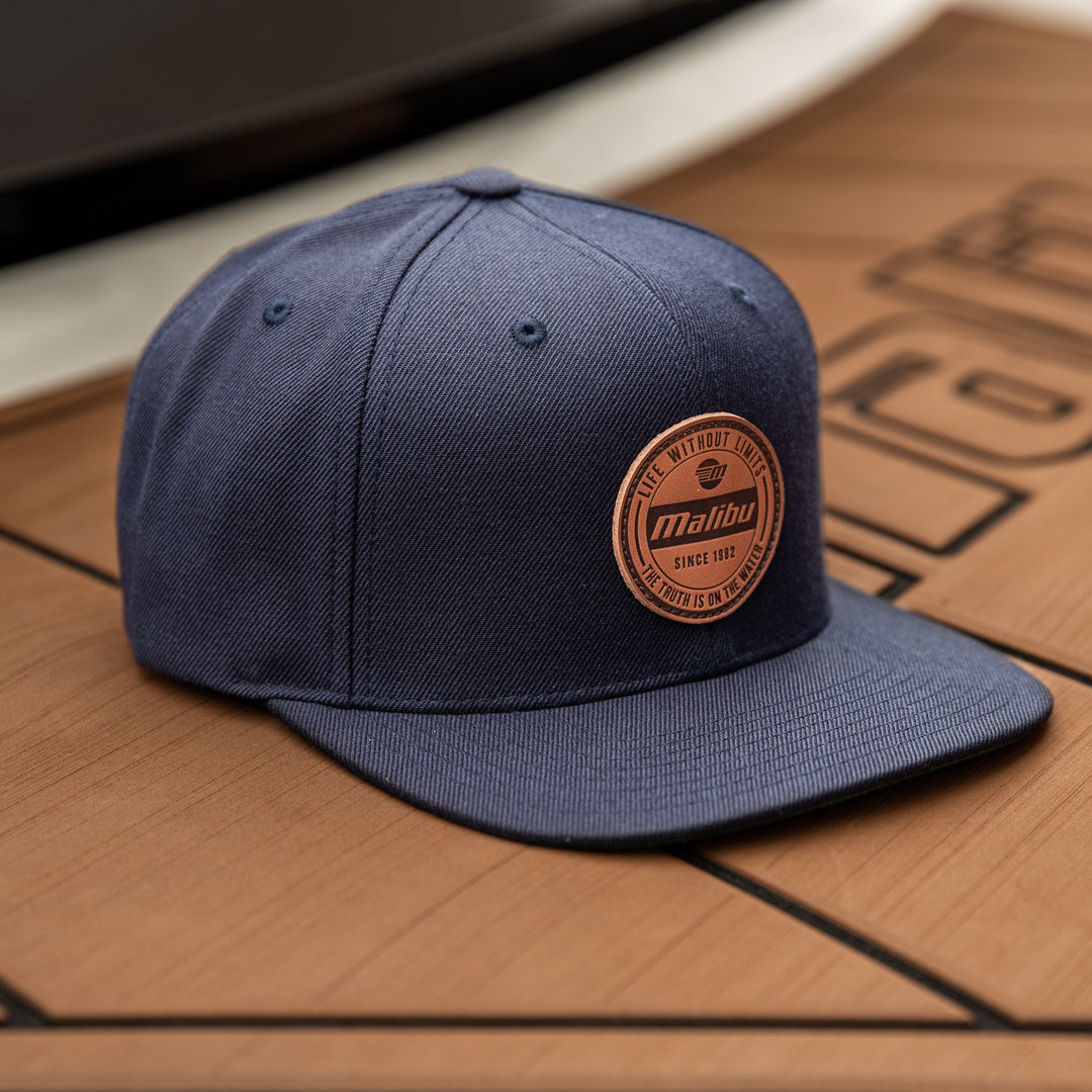 Limitless Hat with a round leather patch logo, navy blue color, and structured brim, placed on a wooden surface.