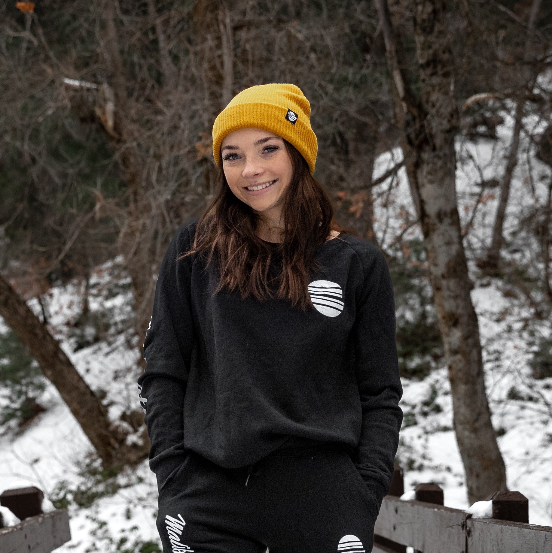 Young woman outdoors in winter wearing a black crewneck sweatshirt with white graphic, yellow beanie, and black pants.